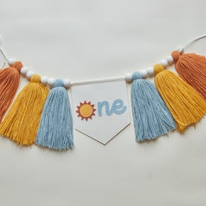 May include: A colorful banner with orange, yellow, and blue tassels, strung with white beads. A white pennant in the center displays the word "one" with a sun graphic, perfect for a first birthday celebration.