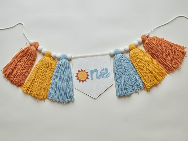 May include: A colorful banner with orange, yellow, and blue tassels, strung with white beads. A white pennant in the center displays the word "one" with a sun graphic, perfect for a first birthday celebration.