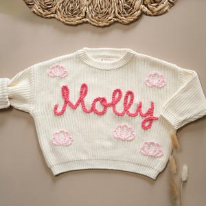 May include: A white knitted sweater with pink embroidered flowers and the name "Molly" in pink yarn.