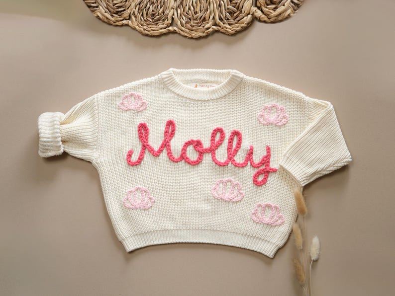 May include: A white knitted sweater with pink embroidered flowers and the name "Molly" in pink yarn.