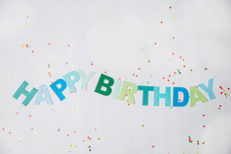 May include: A blue and green felt banner with the words "Happy Birthday" strung on white twine. The banner is surrounded by colorful confetti.