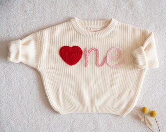 Peut inclure: Un pull en tricot crème à manches longues orné d'un cœur rouge texturé sur le côté gauche et du mot one brodé en écriture cursive rose clair sur le devant. Le pull présente un col rond côtelé et des manches à revers.
