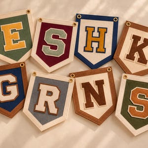 May include: A collection of eight felt pennants featuring the letters E, S, H, K, G, R, N, and S. Each pennant has a cream-colored border, a metal grommet at the top corners, and a large, block-style felt letter centered on a solid-colored background.