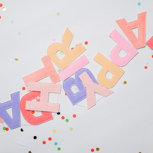 May include: A colorful felt banner spelling out "Happy Birthday" with pastel pink, orange, purple, and red letters. The banner is strung with white thread and is surrounded by confetti.
