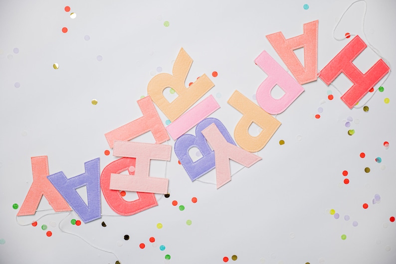 May include: A colorful felt banner spelling out "Happy Birthday" with pastel pink, orange, purple, and red letters. The banner is strung with white thread and is surrounded by confetti.