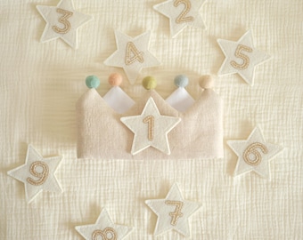 May include: A beige fabric crown featuring a central white star with the number 1 embroidered in gold thread. The crown is topped with small pastel-colored felt balls and is surrounded by several white star-shaped patches numbered 2 through 9.