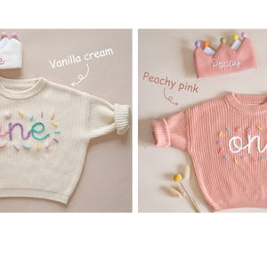 May include: Two knitted baby sweaters with the word "one" embroidered on the front in colorful thread. The sweaters are in vanilla cream and peachy pink colors. Each sweater is paired with a crown headband, one with the name "Rosie" and the other with the name "Poppy".
