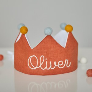 May include: A burnt orange fabric crown with white trim and the name "Oliver" embroidered in white. The crown is decorated with yellow and light blue pom-poms. Scattered around the crown are small, colorful felt balls.