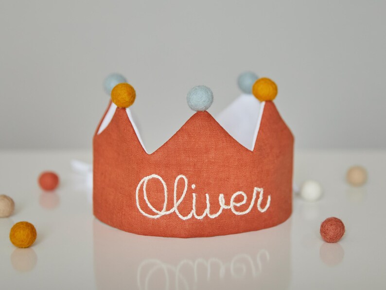 May include: A burnt orange fabric crown with white trim and the name "Oliver" embroidered in white. The crown is decorated with yellow and light blue pom-poms. Scattered around the crown are small, colorful felt balls.