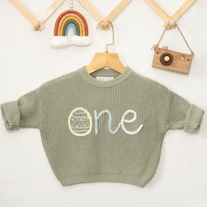 K&ouml;nnte beinhalten: Ein salbeigr&uuml;ner Strickpullover mit einem wei&szlig;en, blauen und gelben gestickten "eins" und einem gelben Ei-Design.