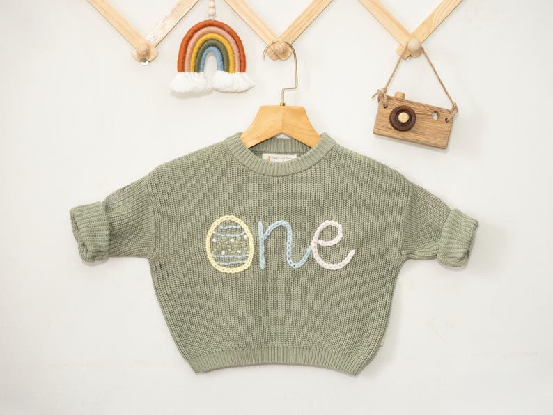 K&ouml;nnte beinhalten: Ein salbeigr&uuml;ner Strickpullover mit einem wei&szlig;en, blauen und gelben gestickten "eins" und einem gelben Ei-Design.