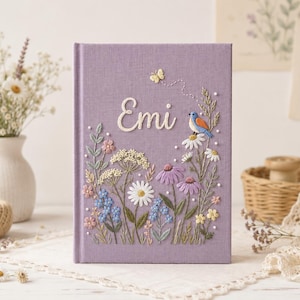 Könnte beinhalten: Ein lavendelfarbenes Hardcover-Buch mit aufwendiger Stickerei auf dem Vorderdeckel. Das Design zeigt eine Vielzahl bunter Wildblumen, einen kleinen blauen Vogel, einen gelben Schmetterling und den Namen Emi in weißem Garn gestickt.