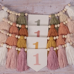 May include: A white felt banner with the number "1" repeated four times in different colors. The banner is decorated with a string of wooden beads and colorful yarn tassels.