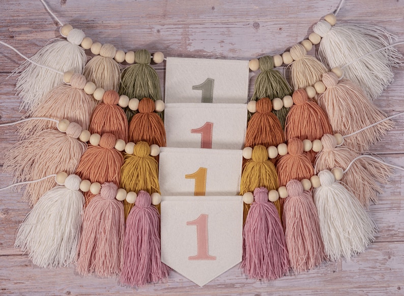 May include: A white felt banner with the number "1" repeated four times in different colors. The banner is decorated with a string of wooden beads and colorful yarn tassels.