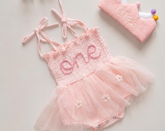 May include: A light pink romper with a smocked bodice featuring the word one in embroidered script, tie-up shoulder straps, and a tulle skirt overlay with small white floral appliqués, paired with a matching pink crown labeled with Mila.