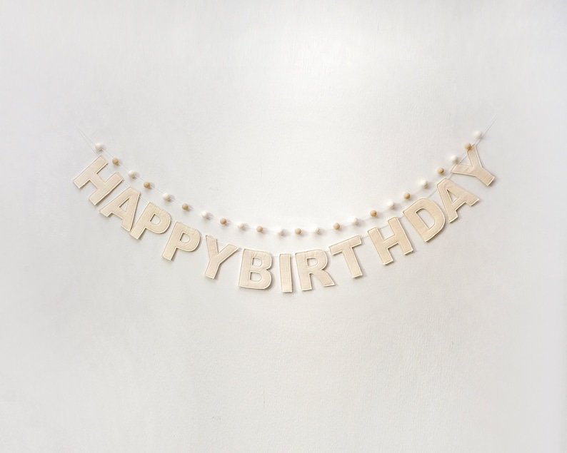 Pode incluir: Um fundo branco com uma faixa bege que diz "Happy Birthday" em letras mai&uacute;sculas. A faixa &eacute; enfiada com pompons brancos e bege.