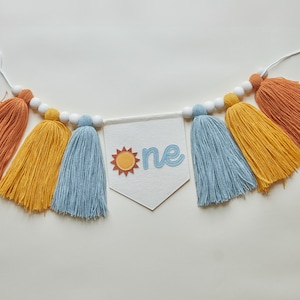 May include: A colorful banner with orange, yellow, and blue tassels, strung with white beads. A white pennant in the center displays the word "one" with a sun graphic, perfect for a first birthday celebration.