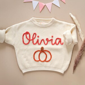May include: A white knitted sweater with the name "Olivia" embroidered in red yarn. The sweater features a pumpkin design in orange and brown yarn.