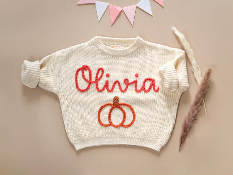 May include: A white knitted sweater with the name "Olivia" embroidered in red yarn. The sweater features a pumpkin design in orange and brown yarn.