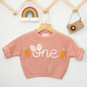 K&ouml;nnte beinhalten: Ein rosa gestrickter Pullover mit einem wei&szlig;en gestickten "eins" und Karotten-Applikationen.