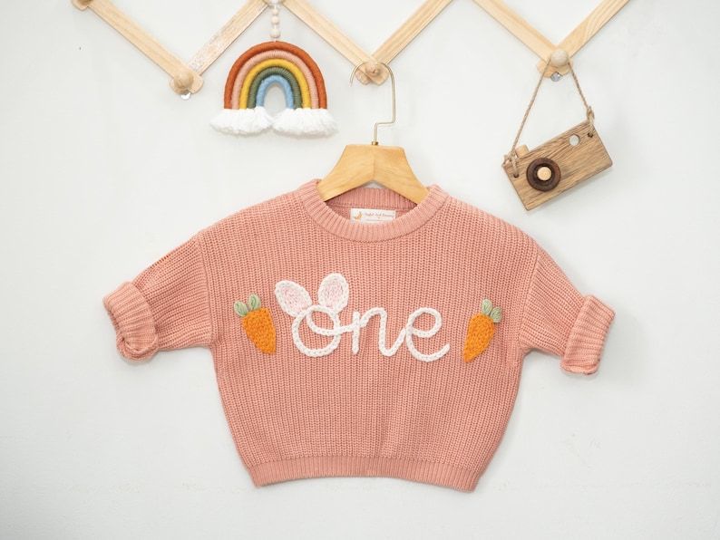 K&ouml;nnte beinhalten: Ein rosa gestrickter Pullover mit einem wei&szlig;en gestickten "eins" und Karotten-Applikationen.