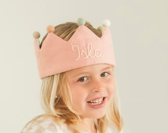 May include: A soft pink fabric crown featuring the name Isla embroidered in white cursive script on the front. The crown is topped with small, round, multi-colored felt pom-poms in shades of beige, sage green, and white along the pointed edges.