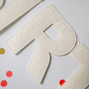 May include: White felt letters spelling out the word "GIRL" with white stitching around the edges. The letters are on a white background with colorful confetti.