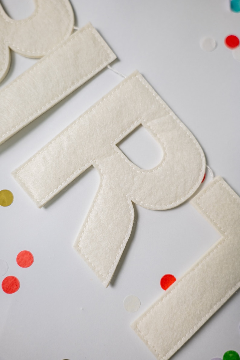 May include: White felt letters spelling out the word "GIRL" with white stitching around the edges. The letters are on a white background with colorful confetti.
