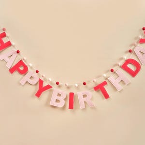 K&ouml;nnte beinhalten: Ein rosa-rotes Filzbanner mit den Worten "Happy Birthday" auf einer wei&szlig;en Schnur mit wei&szlig;en und rosa Pompons.