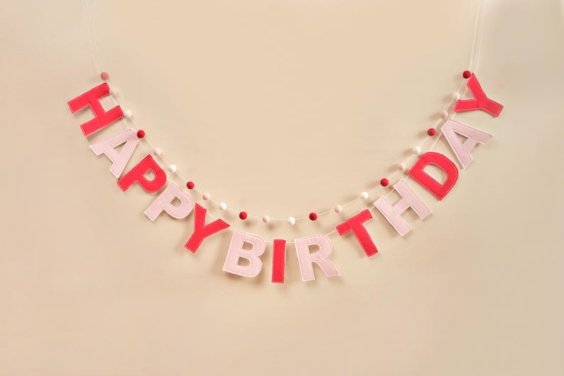 K&ouml;nnte beinhalten: Ein rosa-rotes Filzbanner mit den Worten "Happy Birthday" auf einer wei&szlig;en Schnur mit wei&szlig;en und rosa Pompons.