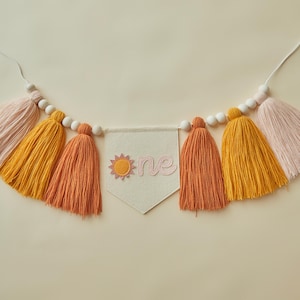 May include: A decorative banner with tassels in shades of pink, orange, and yellow, strung with wooden beads. A small banner in the center displays the word "one" with a sun graphic, perfect for a first birthday celebration.