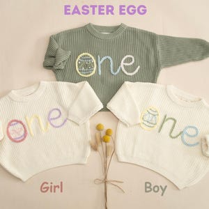 K&ouml;nnte beinhalten: Zwei wei&szlig;e Strickpullover mit dem Wort "one" auf der Vorderseite gestickt. Das Wort "one" ist mit einem gestickten Osterei verziert. Ein Pullover ist f&uuml;r ein M&auml;dchen und der andere f&uuml;r einen Jungen.