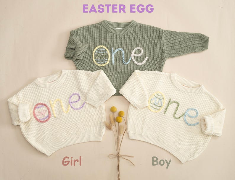 K&ouml;nnte beinhalten: Zwei wei&szlig;e Strickpullover mit dem Wort "one" auf der Vorderseite gestickt. Das Wort "one" ist mit einem gestickten Osterei verziert. Ein Pullover ist f&uuml;r ein M&auml;dchen und der andere f&uuml;r einen Jungen.