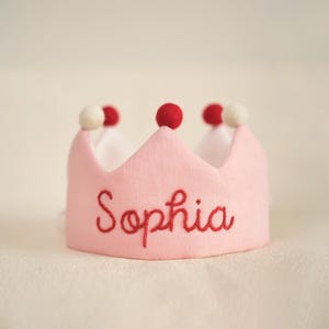 K&ouml;nnte beinhalten: Rosa Filzkrone mit roten Bommeln und dem Namen "Sophia" in roter Farbe gestickt.
