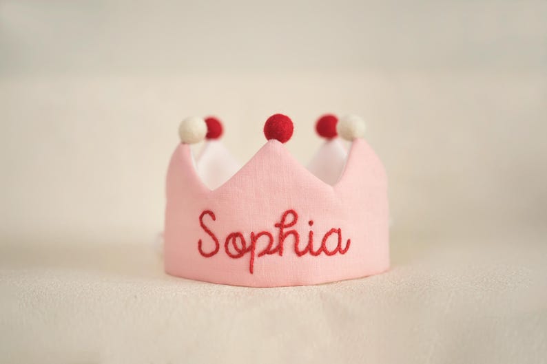 K&ouml;nnte beinhalten: Rosa Filzkrone mit roten Bommeln und dem Namen "Sophia" in roter Farbe gestickt.