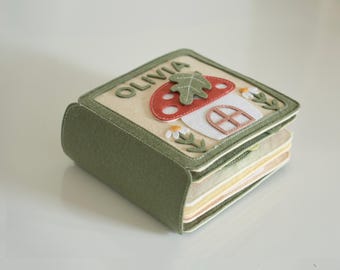 May include: A handmade felt book featuring a mushroom and leaf design. The cover is beige with a red mushroom, white window, and the name "OLIVIA" in green letters. The book's edges are green, and the pages are various pastel colours.