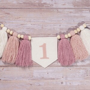 May include: A white banner with the number "1" in pink, decorated with a string of wooden beads and pink and white tassels.
