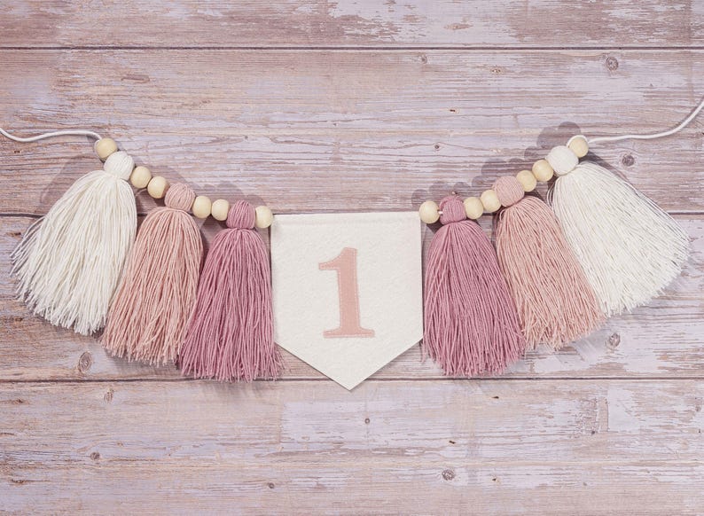 May include: A white banner with the number "1" in pink, decorated with a string of wooden beads and pink and white tassels.