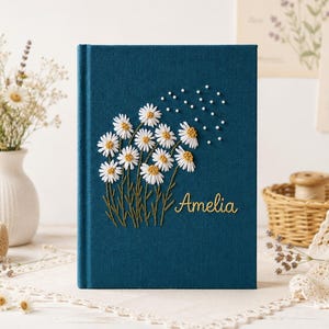 May include: A dark teal fabric-covered book featuring an embroidered design of white daisies with yellow centres and green stems. The name Amelia is embroidered in gold thread on the lower right side, accompanied by small white embroidered dots.