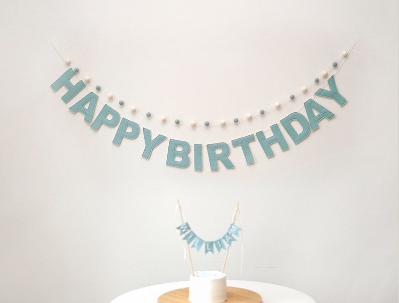 Pode incluir: Uma faixa de feltro azul com as palavras "Happy Birthday" enfiadas com pompons brancos e azuis. Uma faixa de feltro azul menor com a palavra "Liam" est&aacute; em cima de um bolo.