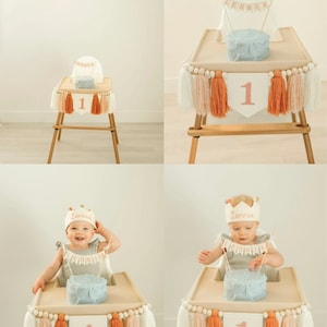 May include: A baby girl sits in a high chair with a blue and white cake in front of her. The high chair is decorated with a white, peach, and orange tassel garland and a banner that says "Lennia". The cake has a number one candle on top. The background is a white wall with a banner that says "Happy Birthday".