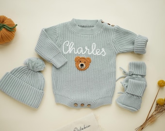May include: A light blue knitted baby outfit set, including a romper with "Charles" embroidered on the front and a teddy bear patch, a beanie with a pom-pom, and matching booties. A small orange pumpkin is also in the image.