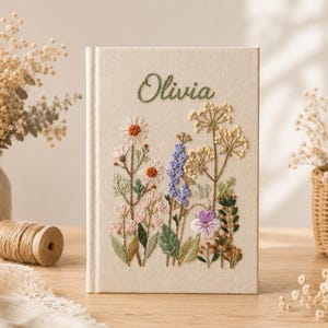 May include: A cream-colored fabric-covered book featuring the embroidered name Olivia in green script at the top. The cover is decorated with a detailed botanical embroidery design showcasing various wildflowers in shades of white, pink, purple, and green.