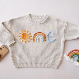 May include: Cream-colored knit sweater with embroidered sun, rainbow, and the word "one" in orange, brown, blue, and light blue. A wooden toy camera and a rainbow-shaped toy are also in the image.