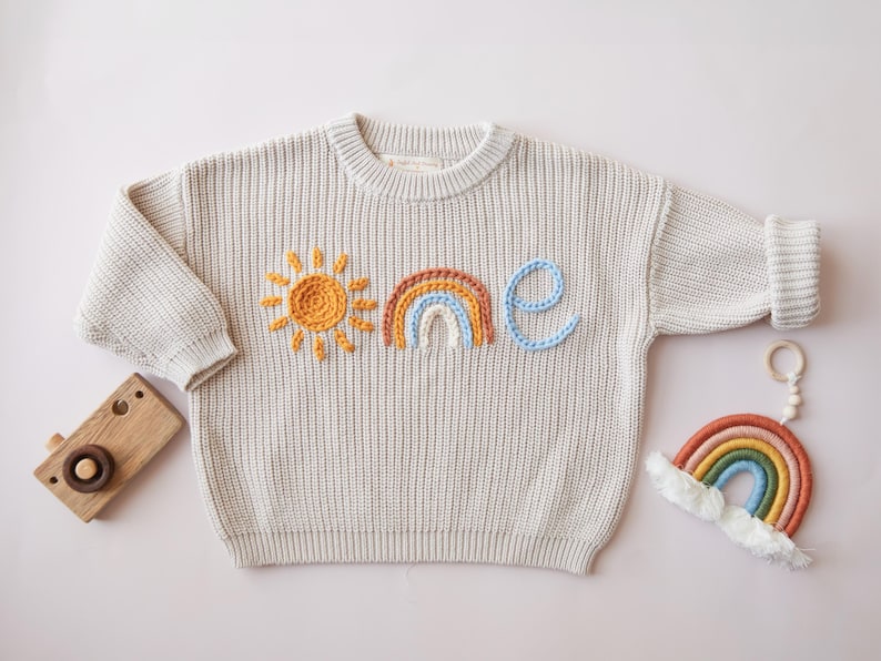 May include: Cream-colored knit sweater with embroidered sun, rainbow, and the word "one" in orange, brown, blue, and light blue. A wooden toy camera and a rainbow-shaped toy are also in the image.