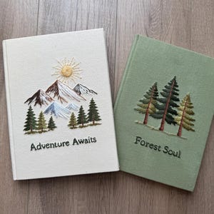 May include: Two hardcover journals with fabric covers. The white journal features embroidered mountains, a yellow sun, green pine trees, and the text "Adventure Awaits." The sage green journal features embroidered pine trees and the text "Forest Soul."