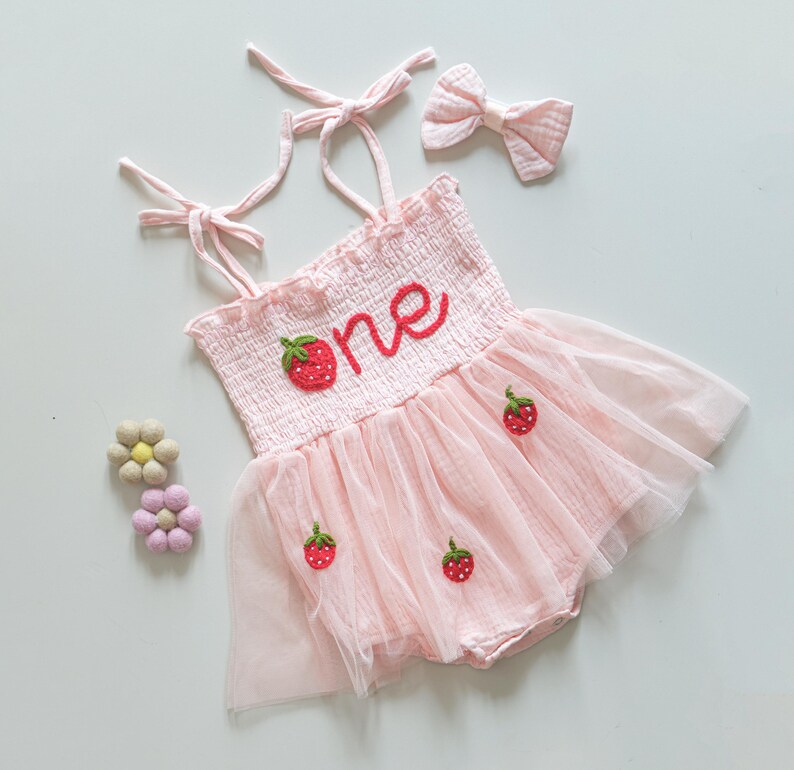 K&ouml;nnte beinhalten: Rosa T&uuml;ll-Babykleid mit einer Erdbeer- und "eins"-Stickerei. Das Kleid hat verstellbare Tr&auml;ger und wird mit einem passenden rosa Stirnband geliefert.
