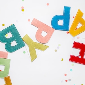 May include: A felt banner with the word "Happy" spelled out in colorful letters. The letters are sewn together with white thread and are arranged in a rainbow pattern. The banner is on a white background with confetti.