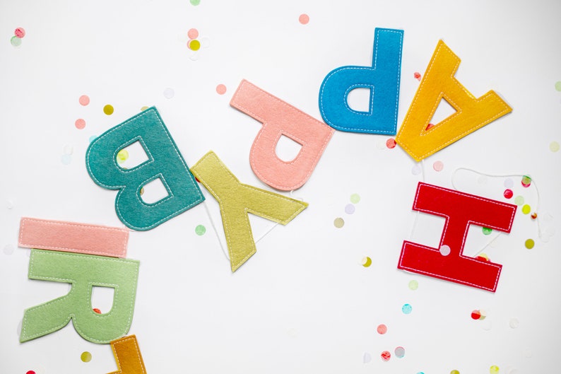May include: A felt banner with the word "Happy" spelled out in colorful letters. The letters are sewn together with white thread and are arranged in a rainbow pattern. The banner is on a white background with confetti.