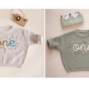 May include: Two knitted baby sweaters with the word "one" embroidered on them. The sweater on the left is oat milk colored and the sweater on the right is dusty olive colored.